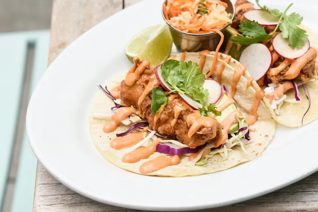 Two fried fish tacos with sauce, cabbage, radish, and cilantro.