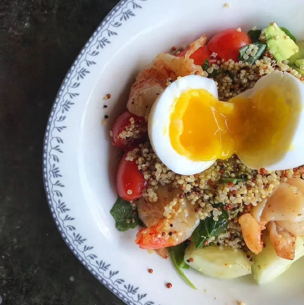 A bowl of quinoa salad with shrimp, tomatoes, and a soft-boiled egg.