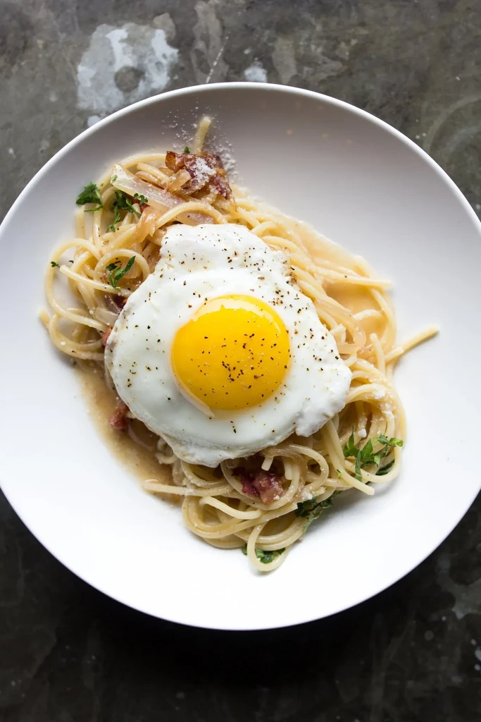 Spaghetti carbonara with a fried egg and black pepper in a white bowl.
