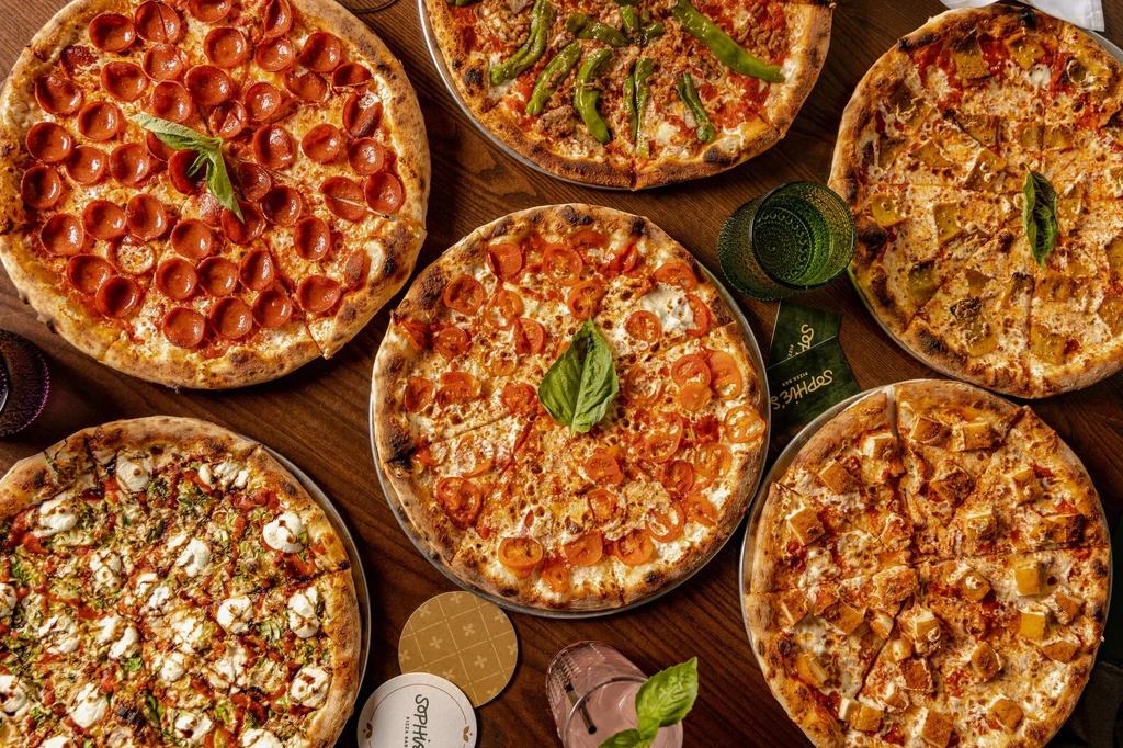 Six different round pizzas with various toppings on a wooden table.