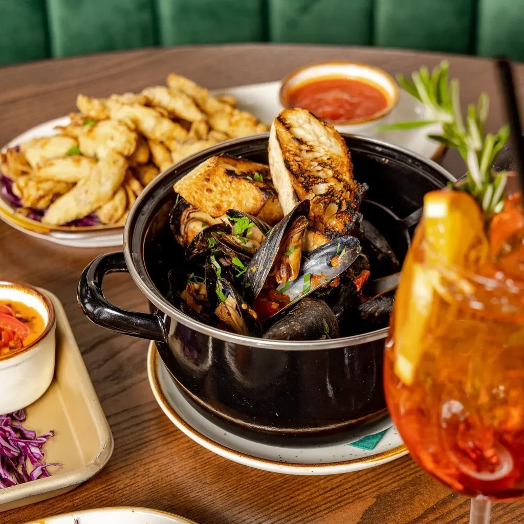 Mussels in a black pot with bread, fries, dips, and an orange drink.