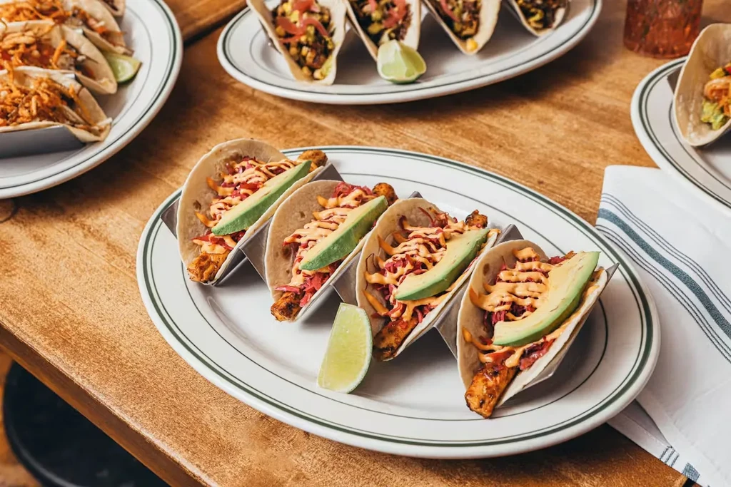 Several plates of tacos with avocado, sauce, and cabbage on a wooden table.