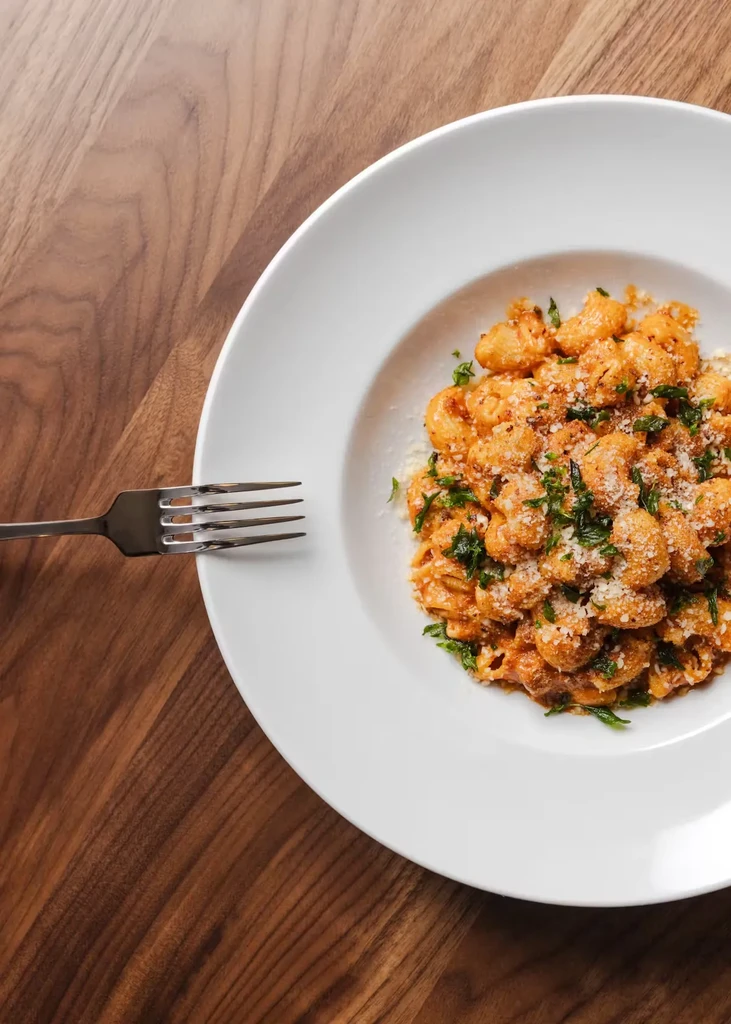 A white bowl of gnocchi with sauce, cheese, and herbs, with a fork.
