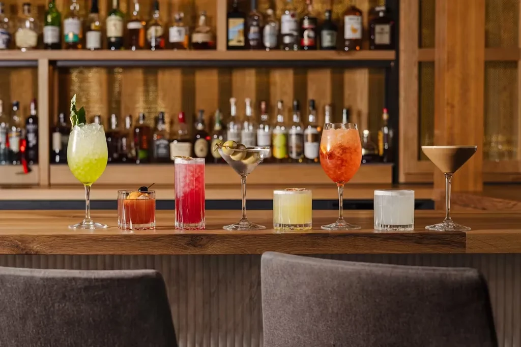Seven cocktails lined up on a bar with liquor bottles in background.