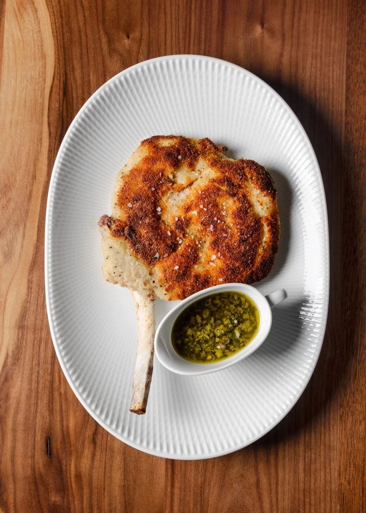 Pan-fried bone-in pork chop with green sauce on white plate.