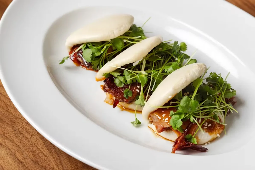 Three open steamed buns with pork belly, cilantro, and microgreens.