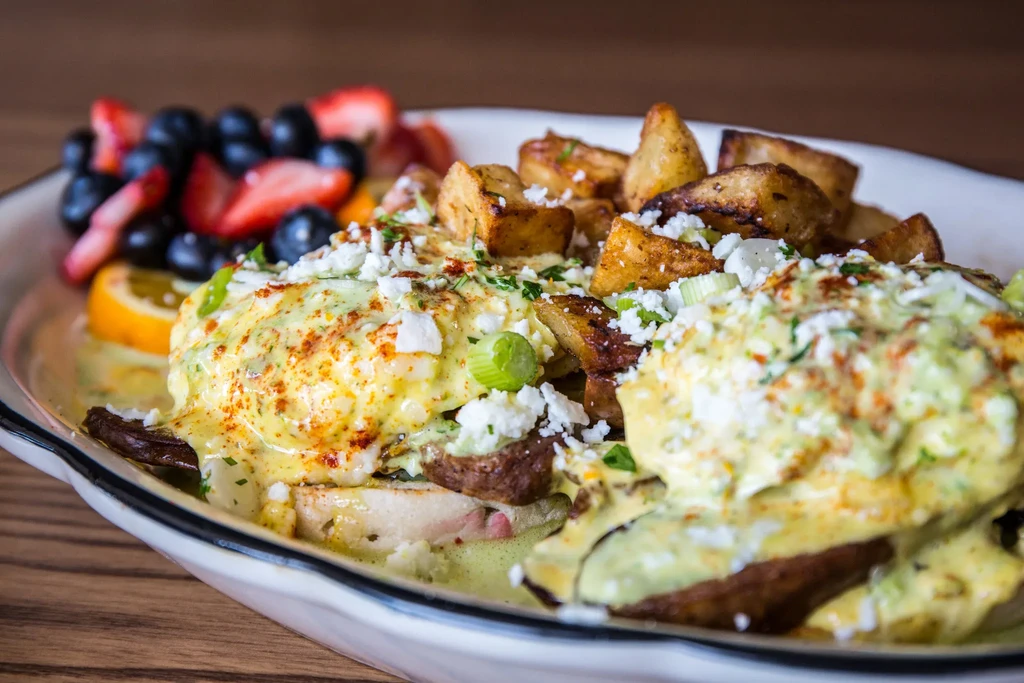 Close-up of Eggs Benedict, potatoes, and fruit on a white plate.