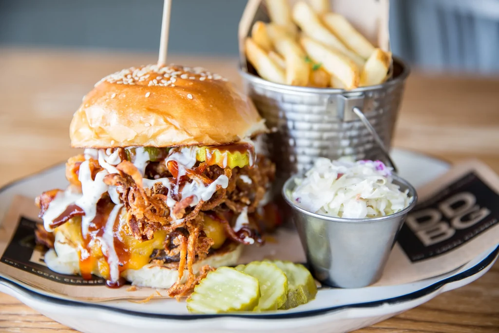 A loaded burger, fries, coleslaw, and pickles on a plate.