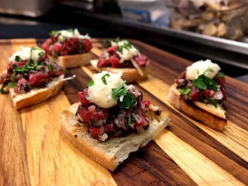 Five beef tartare crostini with cream and herbs on a wooden board.