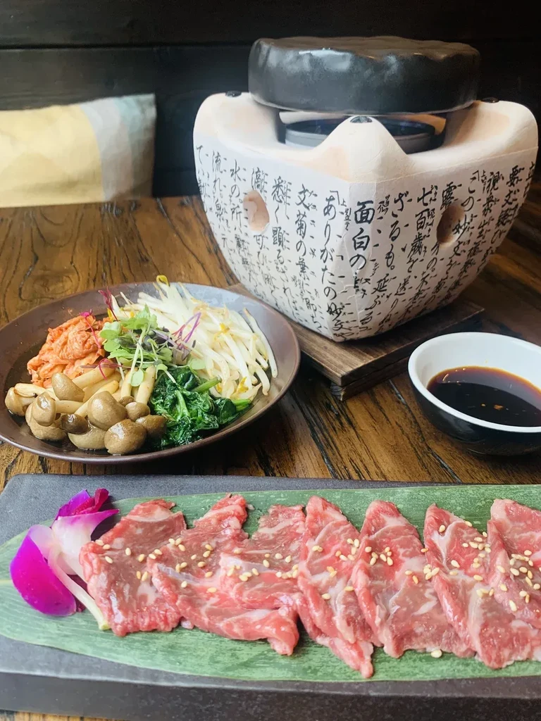 Marbled beef slices, side dishes, and a ceramic grill on a wooden table.