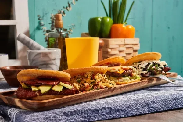 A wooden tray holds five arepas with various fillings on a blue table.