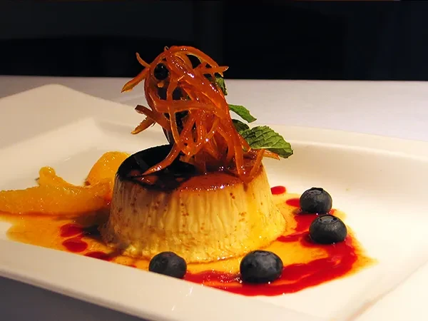 Flan dessert with candied orange peel, blueberries, and mint on a white plate.