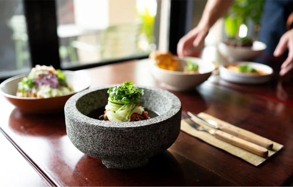 A table set with bowls of food and cutlery, with hands in the background.