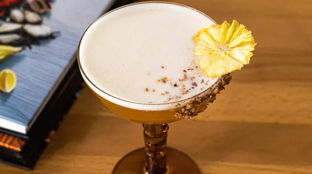 A cocktail with foam, a dried pineapple, and a rim of spices.