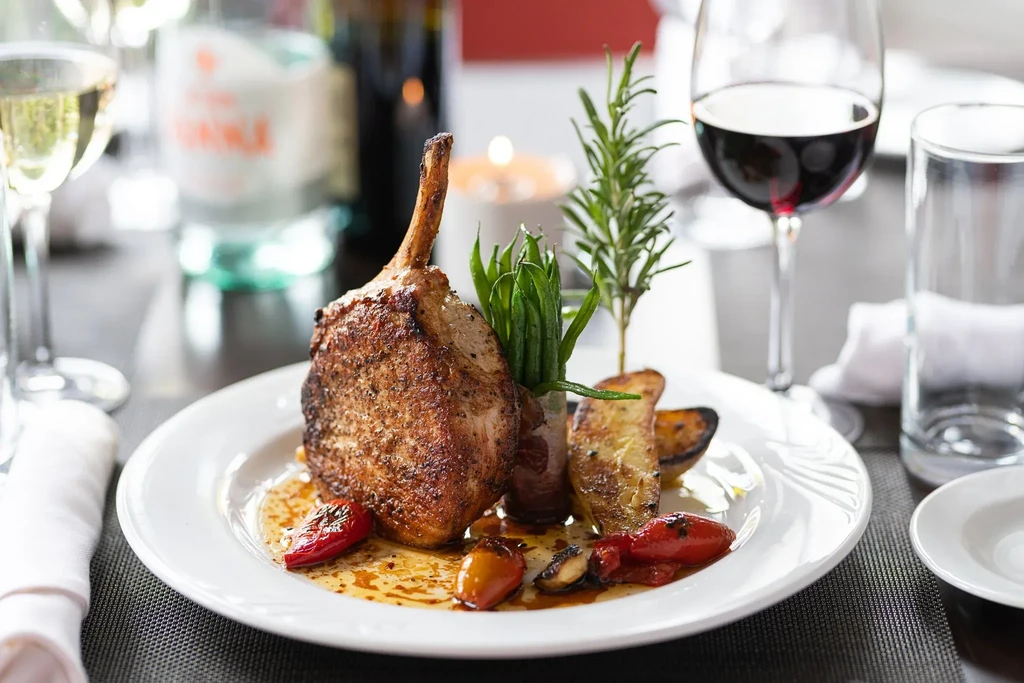 Grilled bone-in pork chop with vegetables, wine, and water at a restaurant.