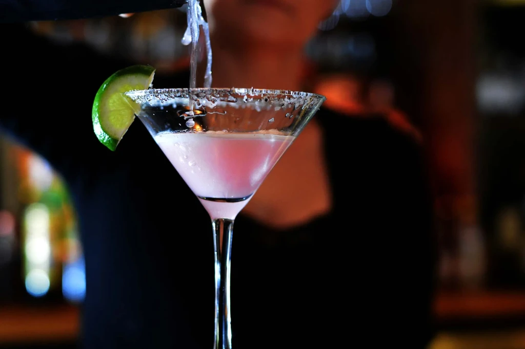 A bartender pours a pink cocktail into a sugar-rimmed martini glass.