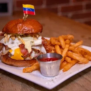 A gourmet burger with a Venezuelan flag, fries, and ketchup on a white plate.