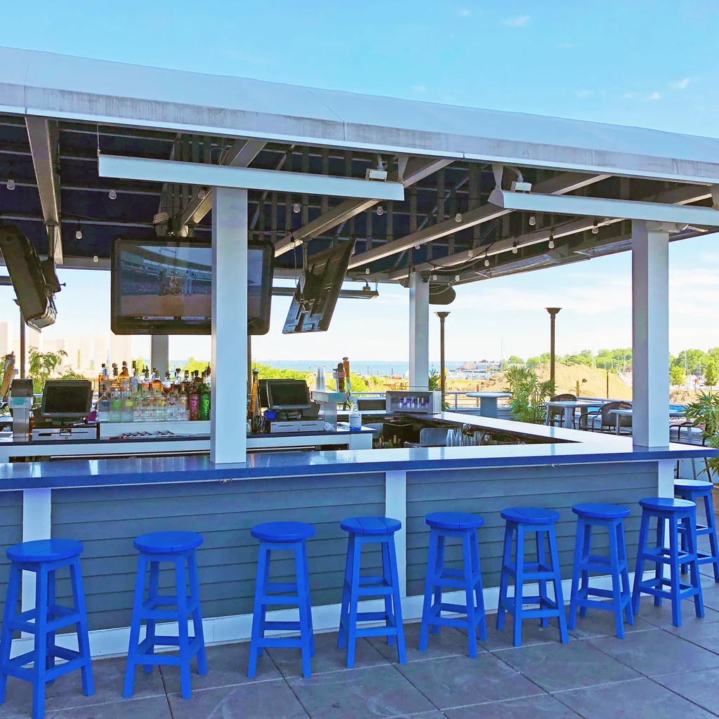 An outdoor bar with blue stools and TVs, overlooking the water.