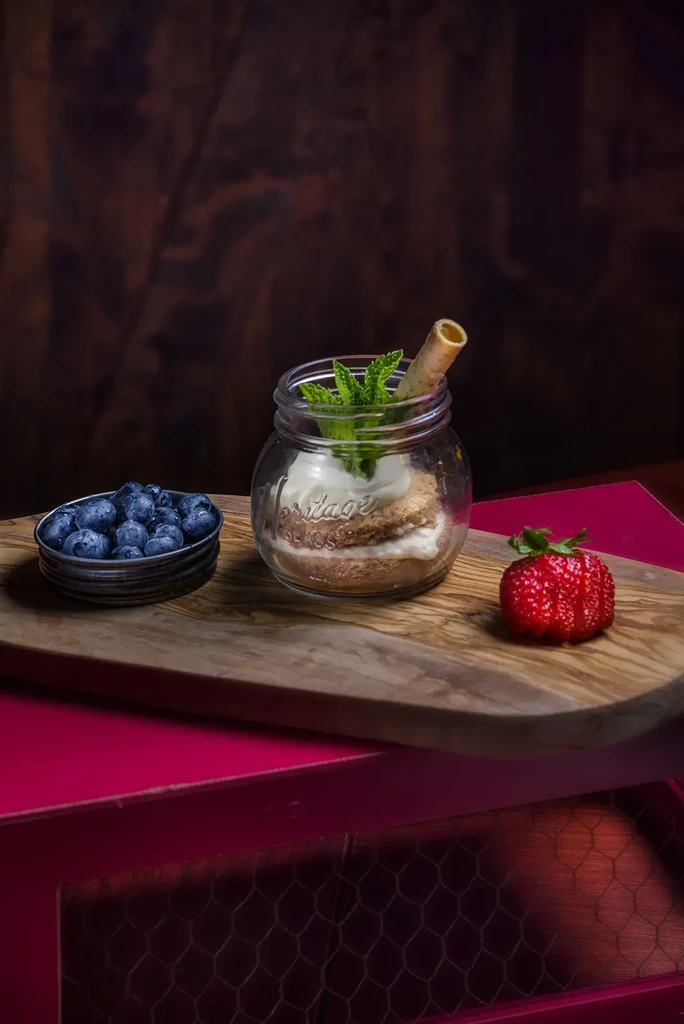 Layered dessert in 'HERITAGE' jar, blueberries, strawberry on wooden board.