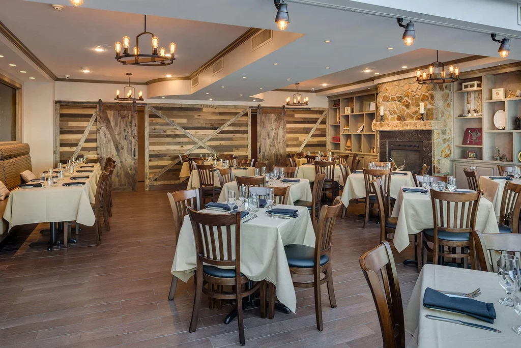 Empty rustic restaurant features stone fireplace, wood walls, set tables.