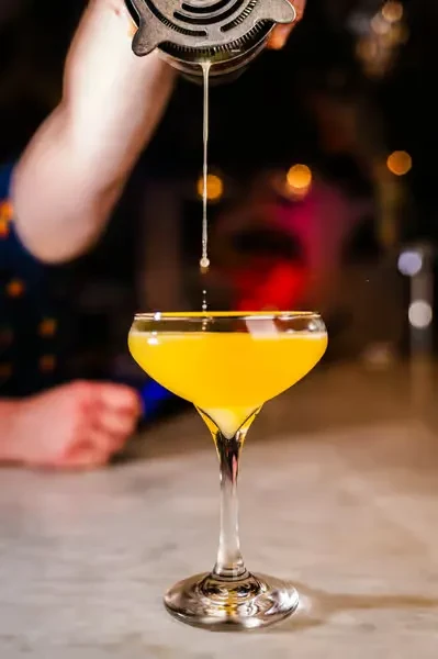 Bartender strains yellow cocktail into a glass on a bar.