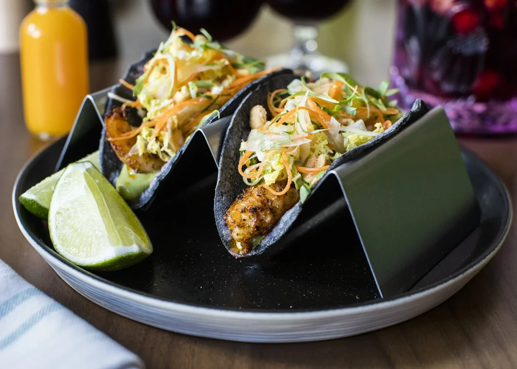 Two black tacos with meat and slaw on a dark plate with lime.