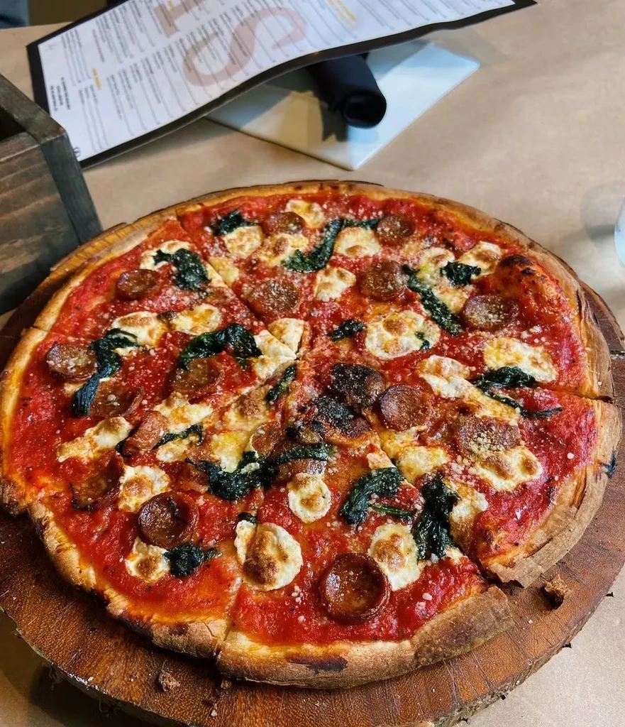 A sliced pizza with pepperoni, spinach, and mozzarella on a wooden board.