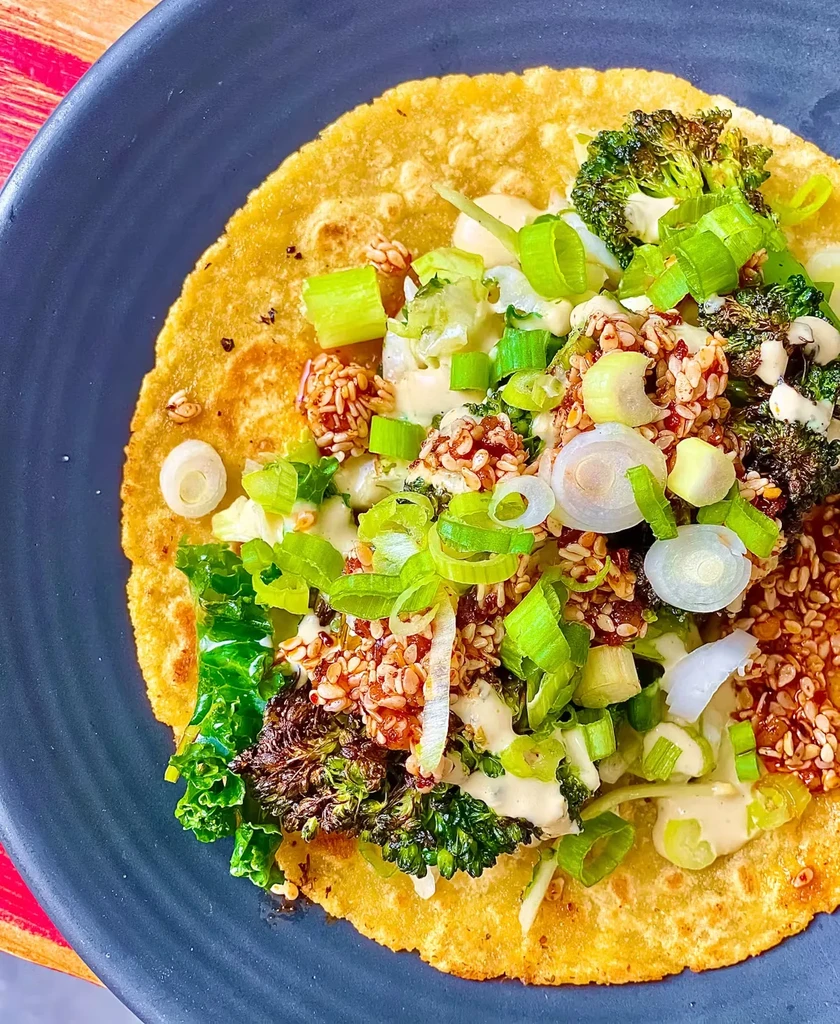 A yellow tortilla with broccoli, seeds, and chopped scallions on a dark plate.