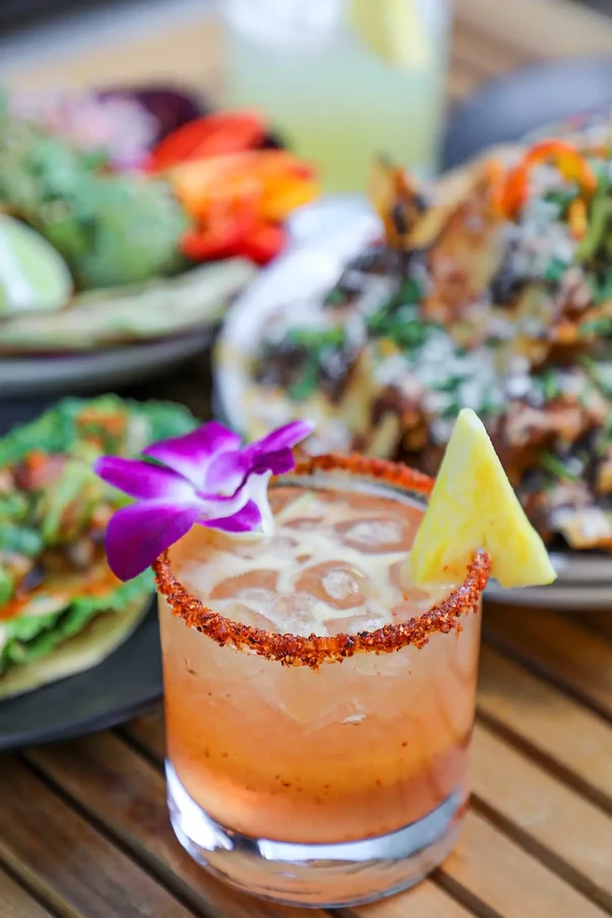 A chili-rimmed cocktail with a purple orchid, pineapple, and blurred food in the background.