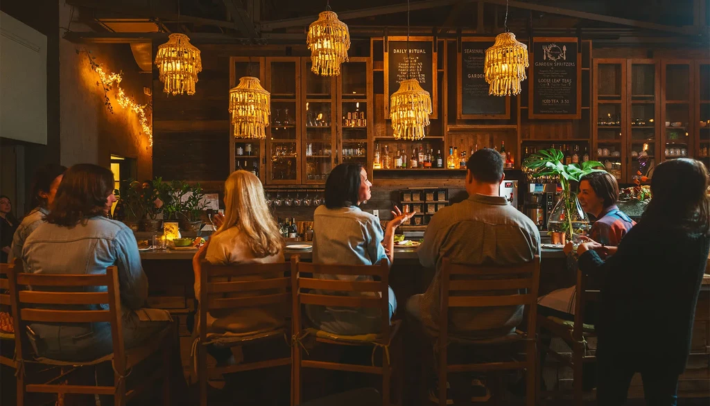 Patrons seated at a bar with woven chandeliers and wooden shelves of bottles.