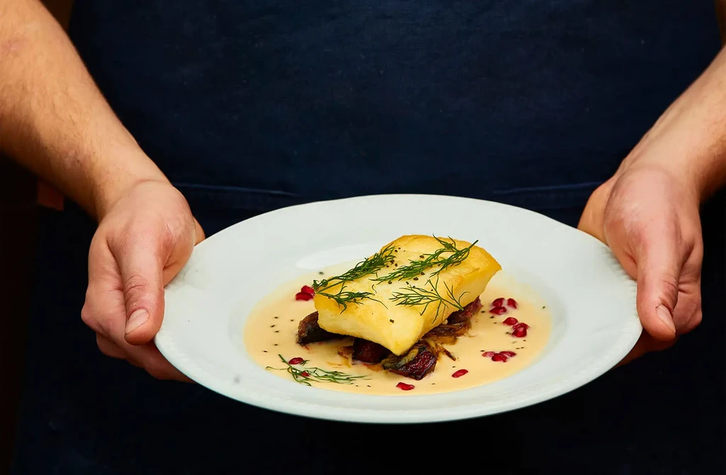 Chef's hands serving a plate of fish and sauce.