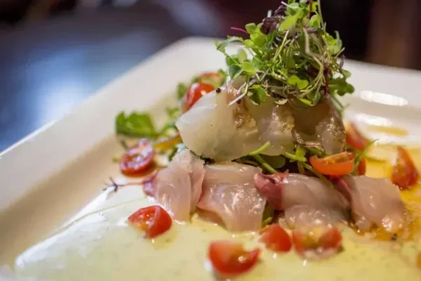 A white rectangular plate with raw fish, tomatoes, greens, and dressing.