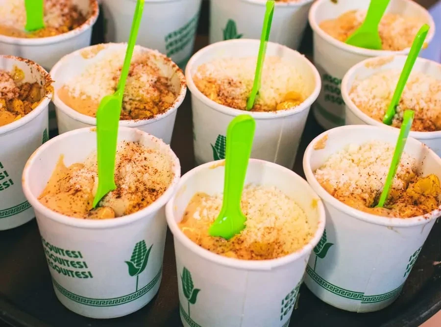 White cups of esquites, topped with cheese and spice, with green spoons.