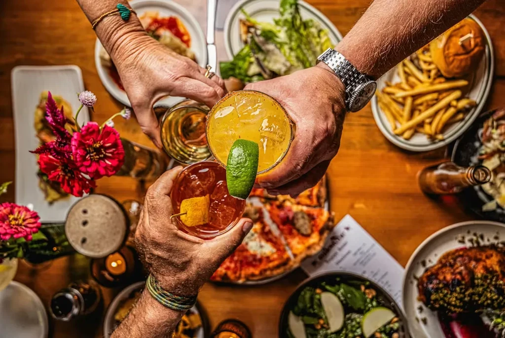 Hands clinking various drinks in a toast over a food-laden table.