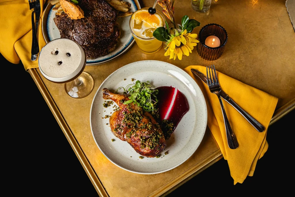 Elegant table with roasted poultry leg, cocktails, steak, and sunflowers.