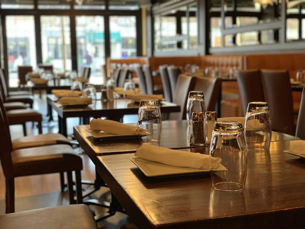 An empty restaurant with set tables, chairs, and large front windows.