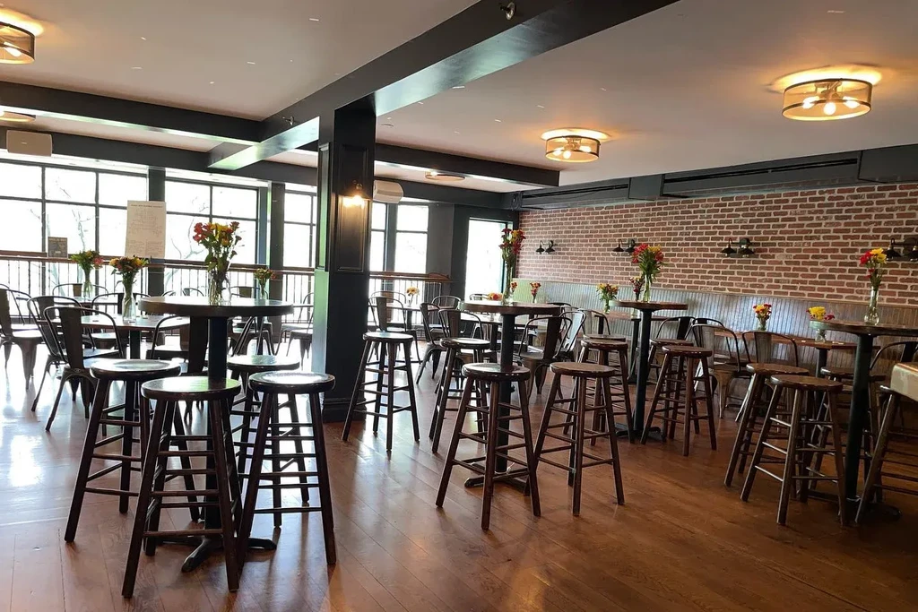 Empty restaurant with high top tables, stools, brick wall, and large windows.