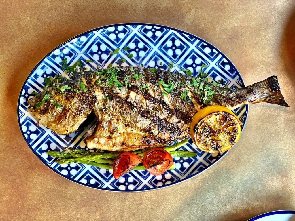 A whole grilled fish with herbs, lemon, asparagus, and tomatoes on a plate.