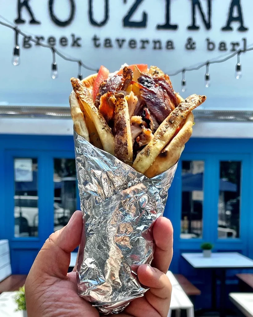 A hand holds a foil-wrapped gyro in front of a blue restaurant.