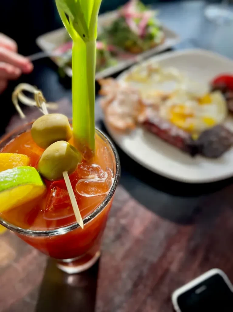 A Bloody Mary cocktail with olives and celery, brunch in the background.
