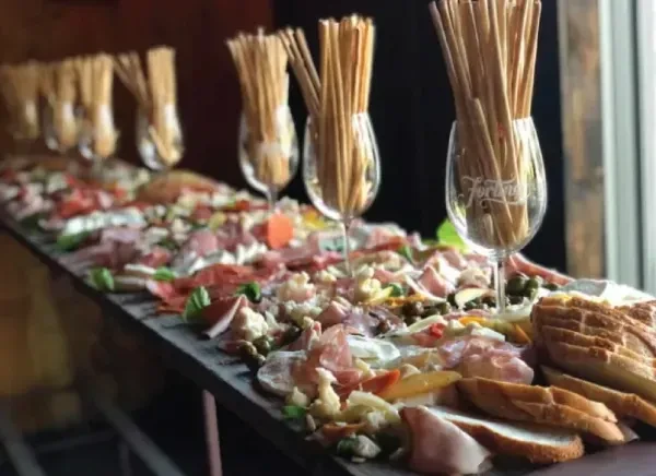 A long table spread with antipasti, bread, and breadsticks in wine glasses.