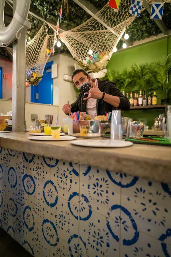 Bartender in a patterned face mask makes drinks behind a tiled bar.