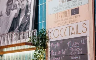 A bar menu lists cocktail prices, with glasses stacked on a shelf.
