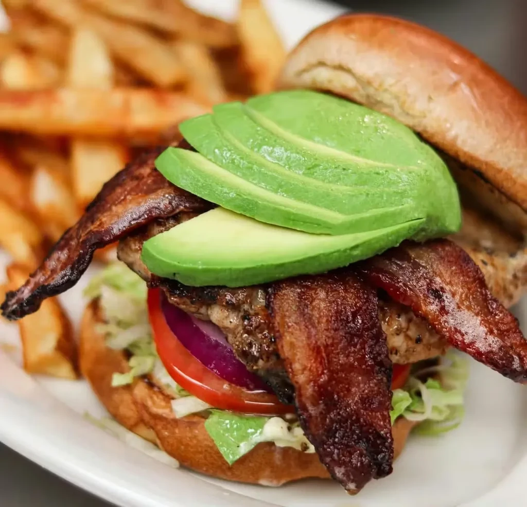 Grilled patty sandwich with bacon, avocado, and blurred french fries.