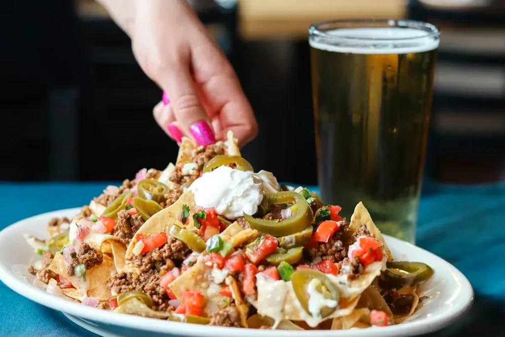 Close-up: hand grabs nacho from beef nachos with toppings and beer.