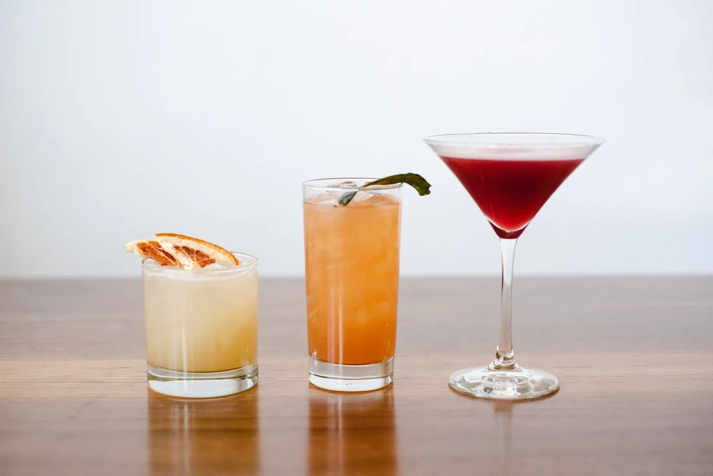 Three distinct colorful cocktails on polished wood against white.