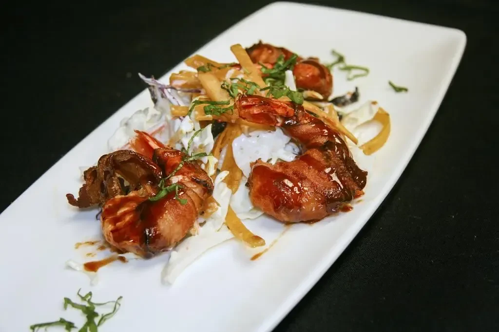 Glazed bacon-wrapped shrimp over cabbage and crispy strips, garnished with herbs.