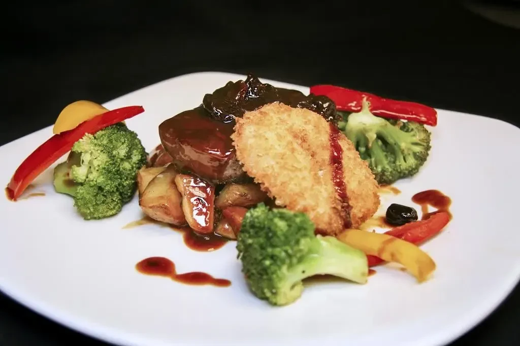 Meticulously plated meal with sauced cutlet, breaded item, and vegetables.