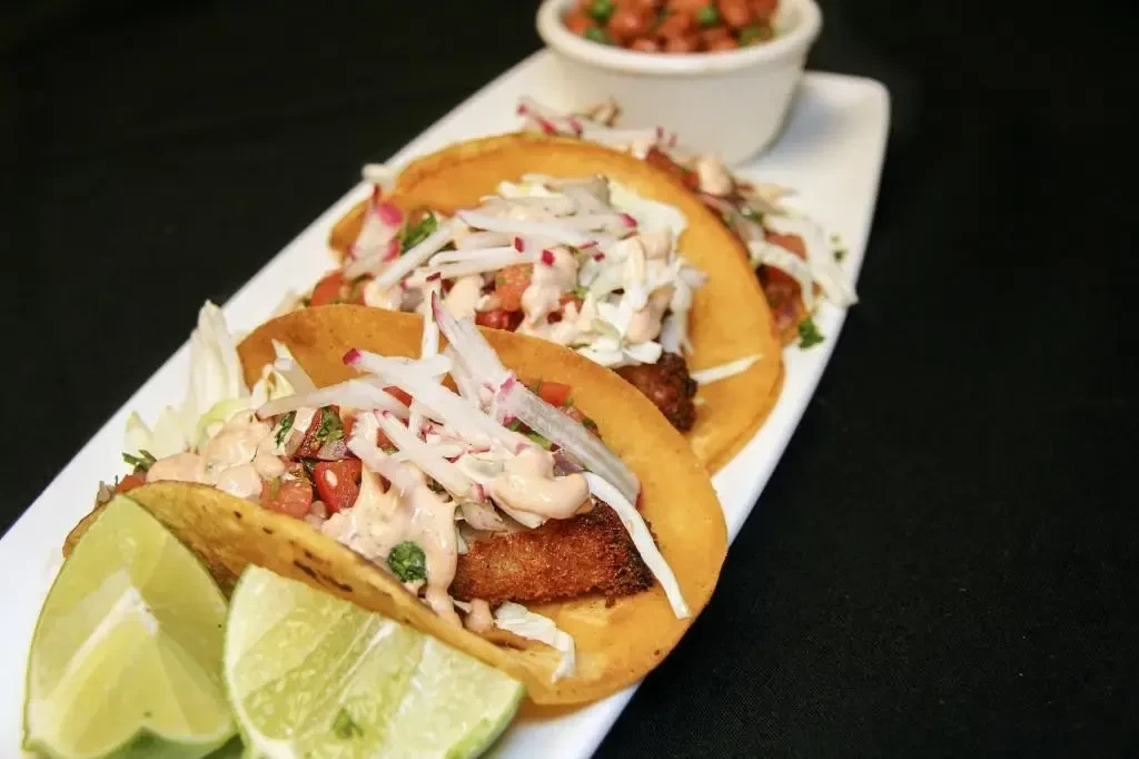 Three crispy tacos with toppings and lime wedges on a white dish.