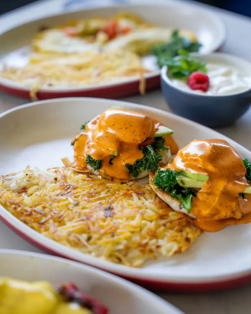 Plate of Eggs Benedict with avocado, hash browns, and raspberry mint side.